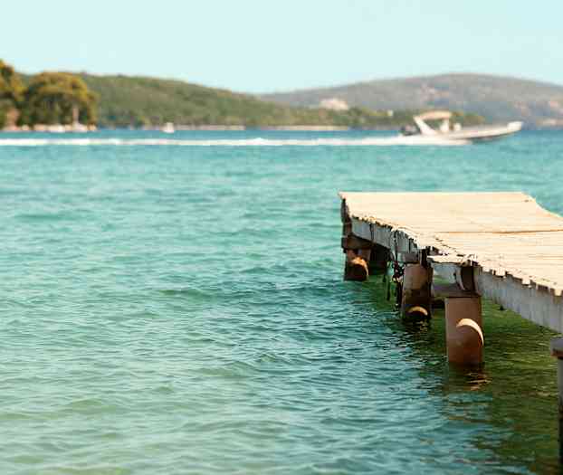 Träbrygga som sträcker sig ut i klart vatten, med en motorbåt och grönskande kullar i bakgrunden. (AI-genererad text)