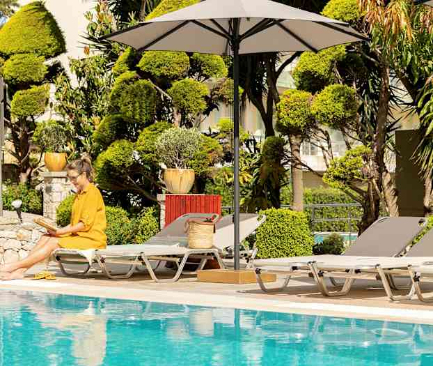 En kvinna i gul klänning sitter med en öppen bok vid en pool, omgiven av solstolar och parasoller. (AI-genererad text)