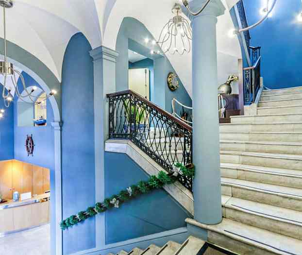 En interiörbild av en hall med en trappa i ljus marmor och blå väggar, dekorerad med gröna girlander. Trappan har räcken med svart metall och trädetaljer. (AI-genererad text)