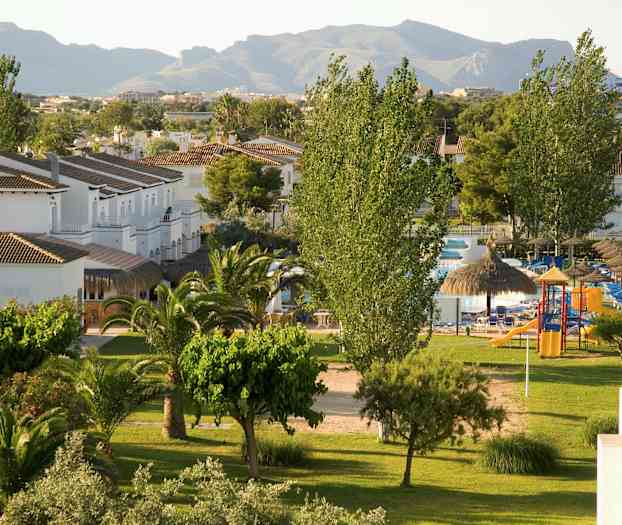 Vy över ett grönområde med träd, gräsmatta och små vita byggnader samt bakgrund med berg. (AI-genererad text)