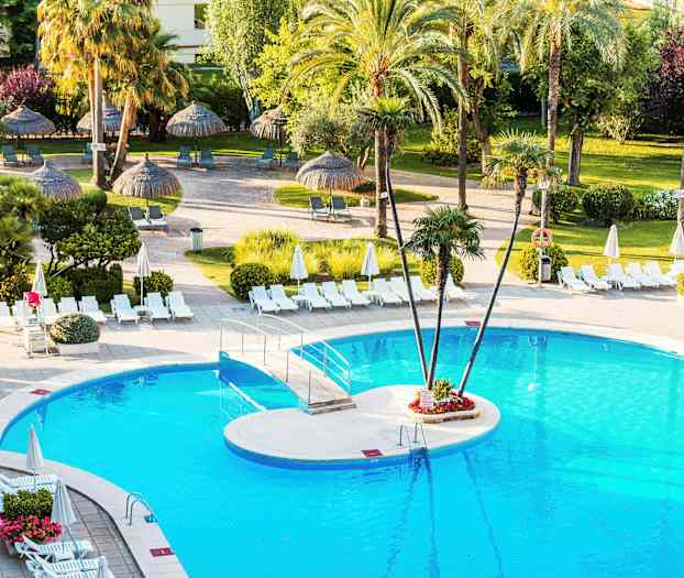 En pool omgiven av solstolar och parasoller, med palmer och grön vegetation i bakgrunden. (AI-genererad text)