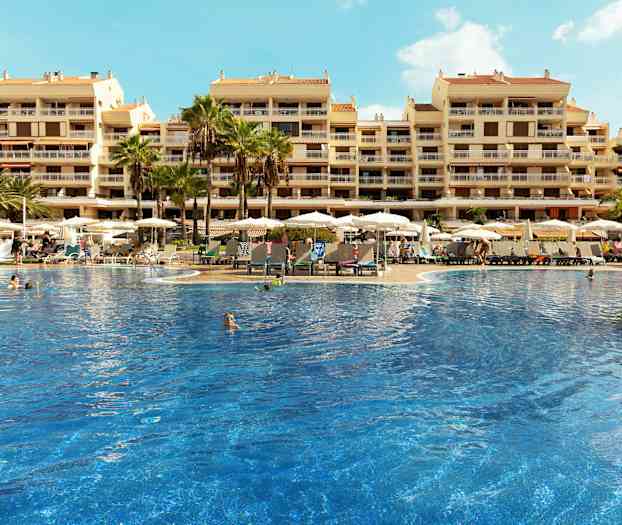 En stor pool omgiven av solstolar och parasoller framför en hotellbyggnad med flera våningar. (AI-genererad text)