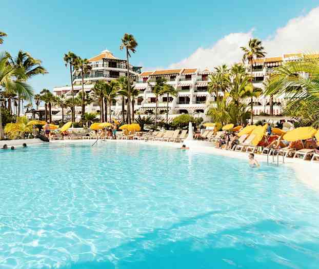 Stor pool omgiven av solstolar och parasoller framför hotellbyggnad. (AI-genererad text)