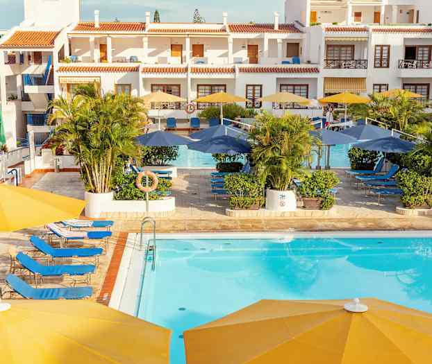 Hotellets poolområde med solstolar, parasoller och byggnader i bakgrunden. (AI-genererad text)