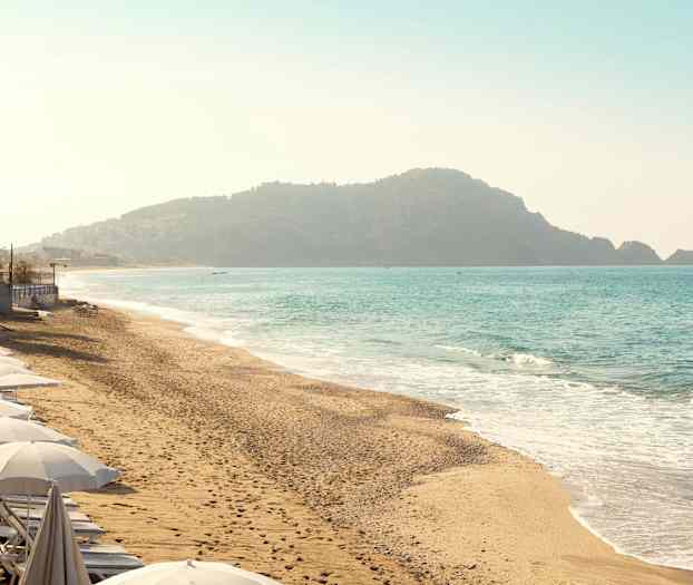 Strand med parasoller längs kustlinjen och bakgrund med berg vid havet. (AI-genererad text)