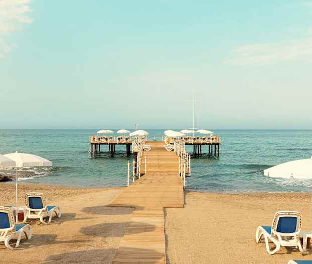 Sandstrand med solstolar och parasoller, en brygga som leder ut till en plattform över vattnet. (AI-genererad text)