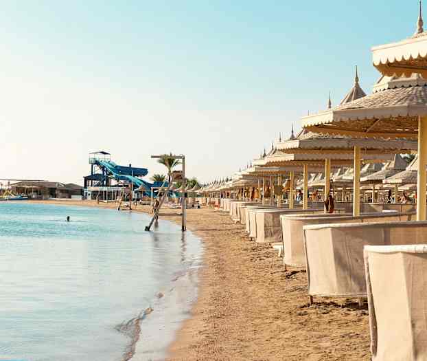 En sandstrand med rad av solparasoller och cabanas intill havet med en vattenrutschbana i bakgrunden. (AI-genererad text)