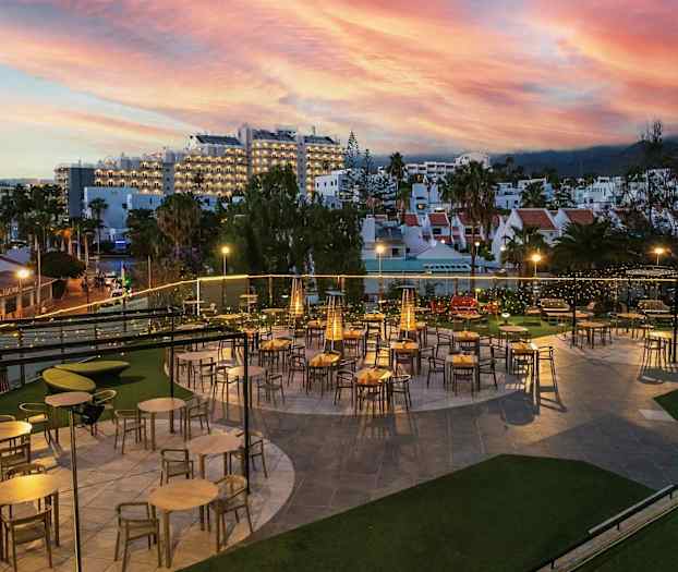 En utomhusrestaurang med bord och stolar under kvällsbelysning. I bakgrunden syns byggnader och en färgstark solnedgång. (AI-genererad text)