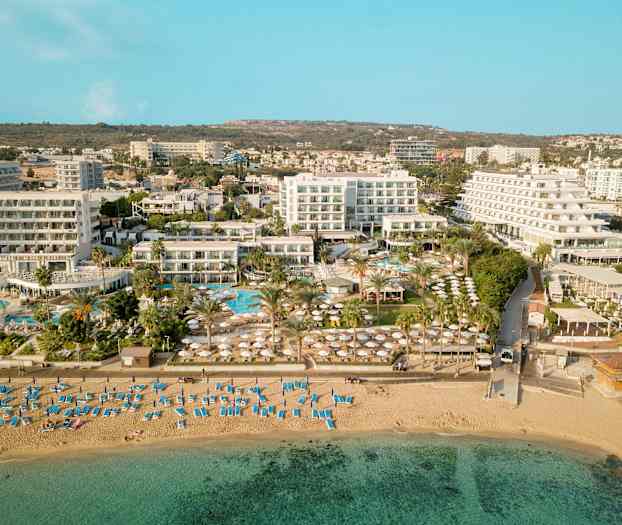 Flygfoto som visar en strand med solstolar och parasoller, samt hotellbyggnader och poolområde i bakgrunden. (AI-genererad text)