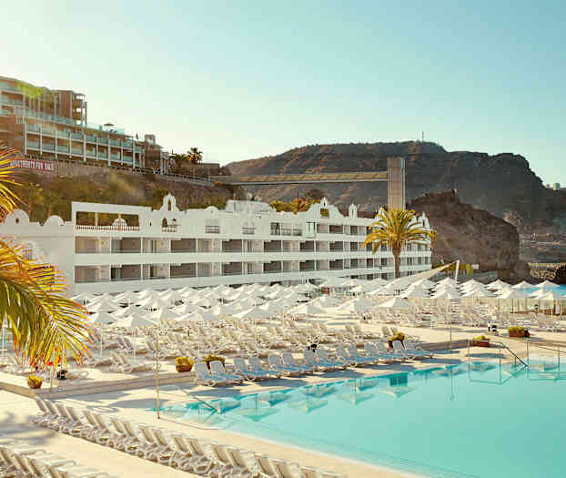 Hotellbyggnad med vita fasader, solstolar och parasoller vid en stor pool, samt klippformationer i bakgrunden. (AI-genererad text)