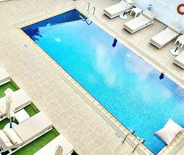 Rektangulär pool med blått vatten omgiven av solstolar och parasoller. (AI-genererad text)