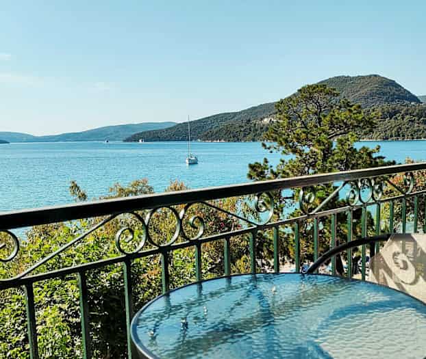 Ett glasbord och stolar på en balkong med utsikt mot havet, grönska och berg i bakgrunden. (AI-genererad text)