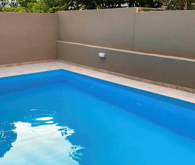 Utomhuspool med klarblått vatten, omgiven av en beige mur och grönska i bakgrunden. (AI-genererad text)