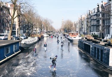 En fryst kanal i Amsterdam, där människor åker skridskor på isen mellan husbåtarna som ligger förtöjda.