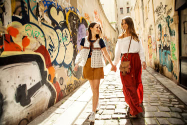 Två unga kvinnor går och håller varandra i handen på en kullerstensgata, vid en graffitimålad mur.