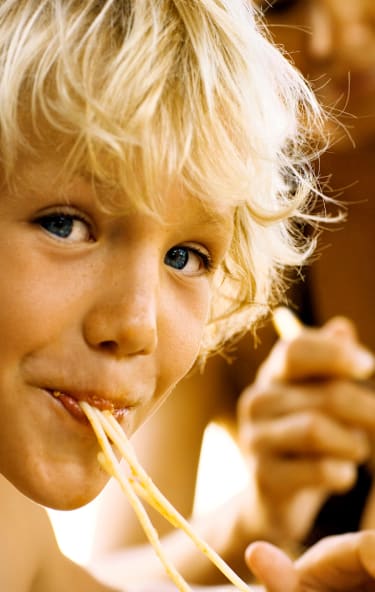 En pojke med blont hår och blåa ögon äter spaghetti samtidigt som han tittar in i kameran.