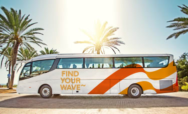 Bild på stor, vit transferbuss som står parkerad framför palmer i ett soligt semsterland.