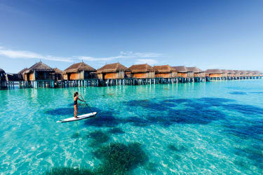 Kvinna som står på en SUP i turkost hav med water villas i bakgrunden.