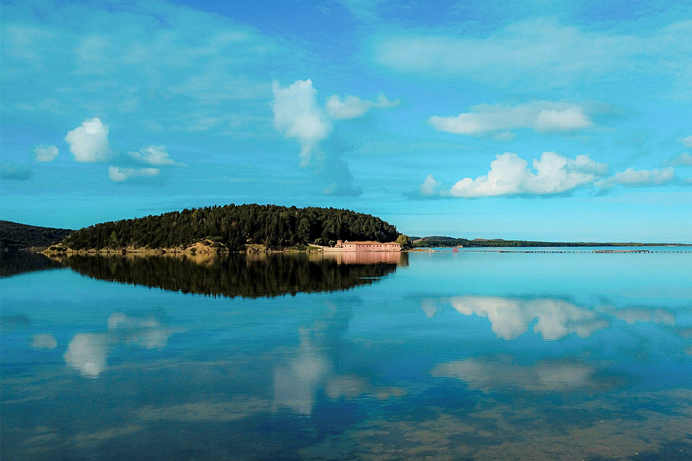 En ö med skog och hus omgiven av stilla vatten, med en klar himmel och moln. (AI-genererad text)