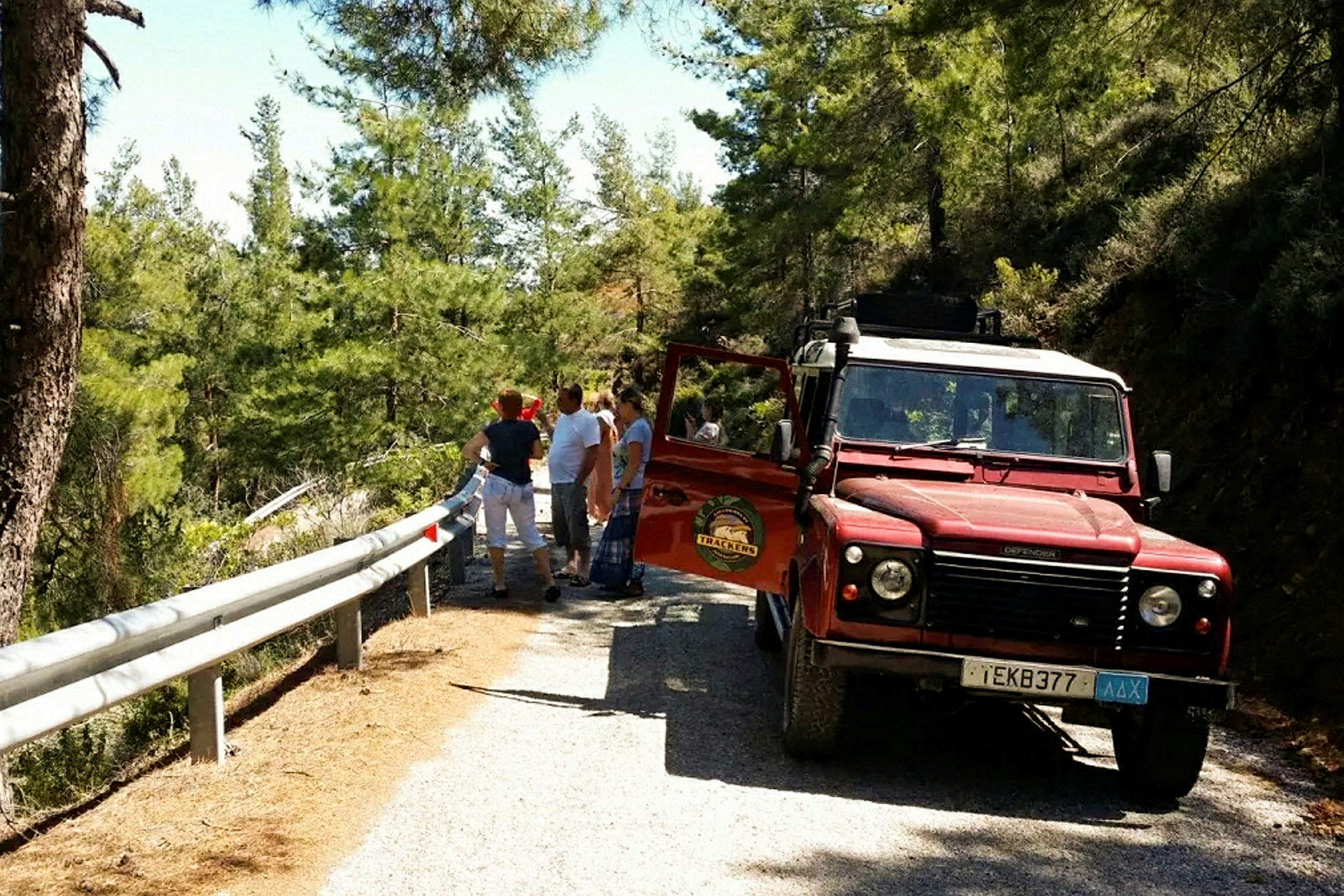 En röd terrängbil står parkerad på en smal väg i skogen, och flera personer står bredvid räcket och pratar. (AI-genererad text)