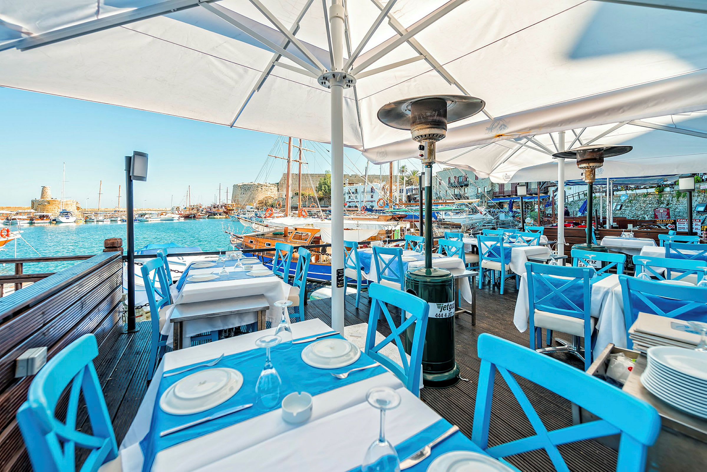 En restaurang med blåa stolar och vita bord vid en hamn. Segelbåtar kan ses i bakgrunden. (AI-genererad text)