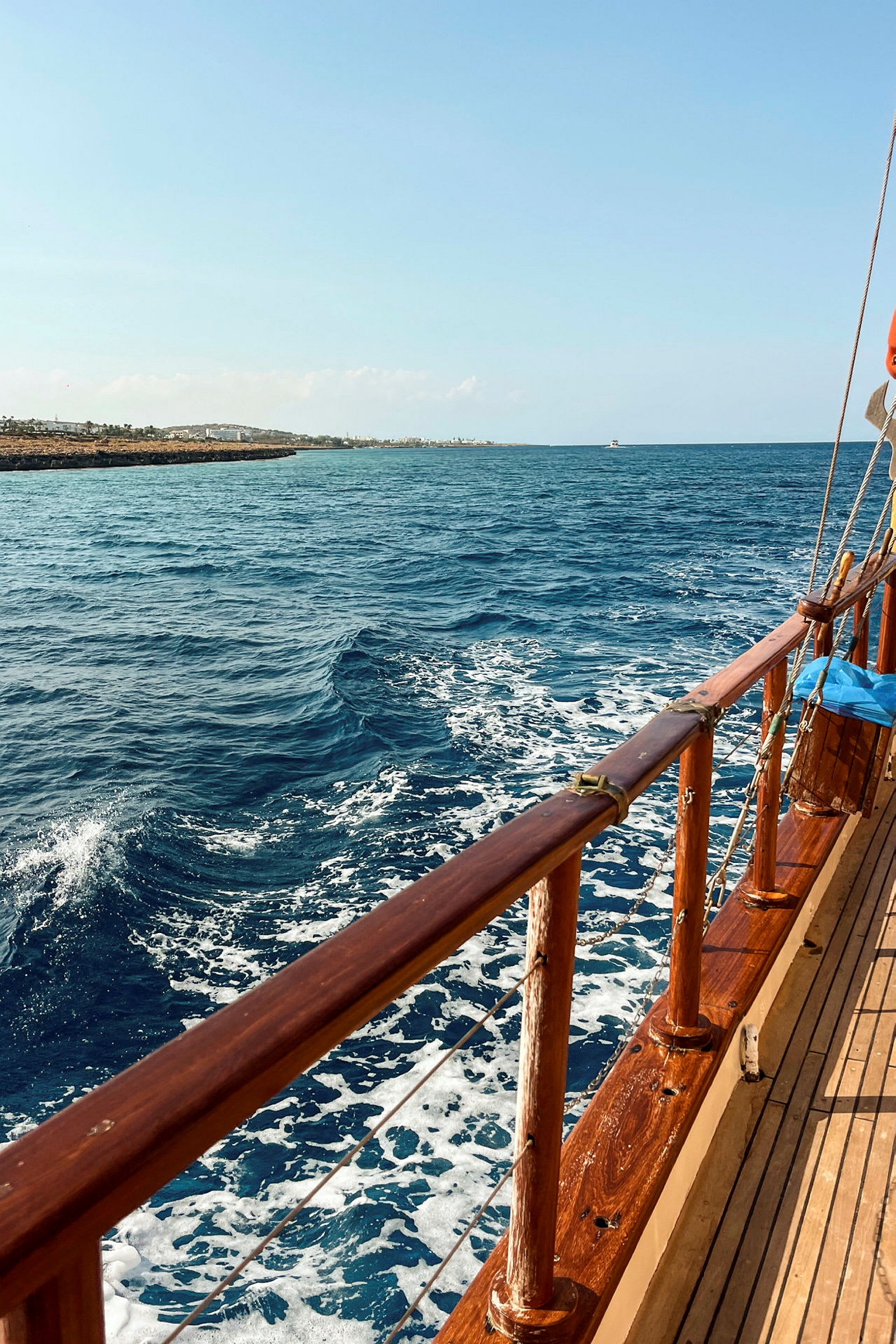 En båt i trä seglar på havet med kustlinjen och byggnader i bakgrunden. (AI-genererad text)