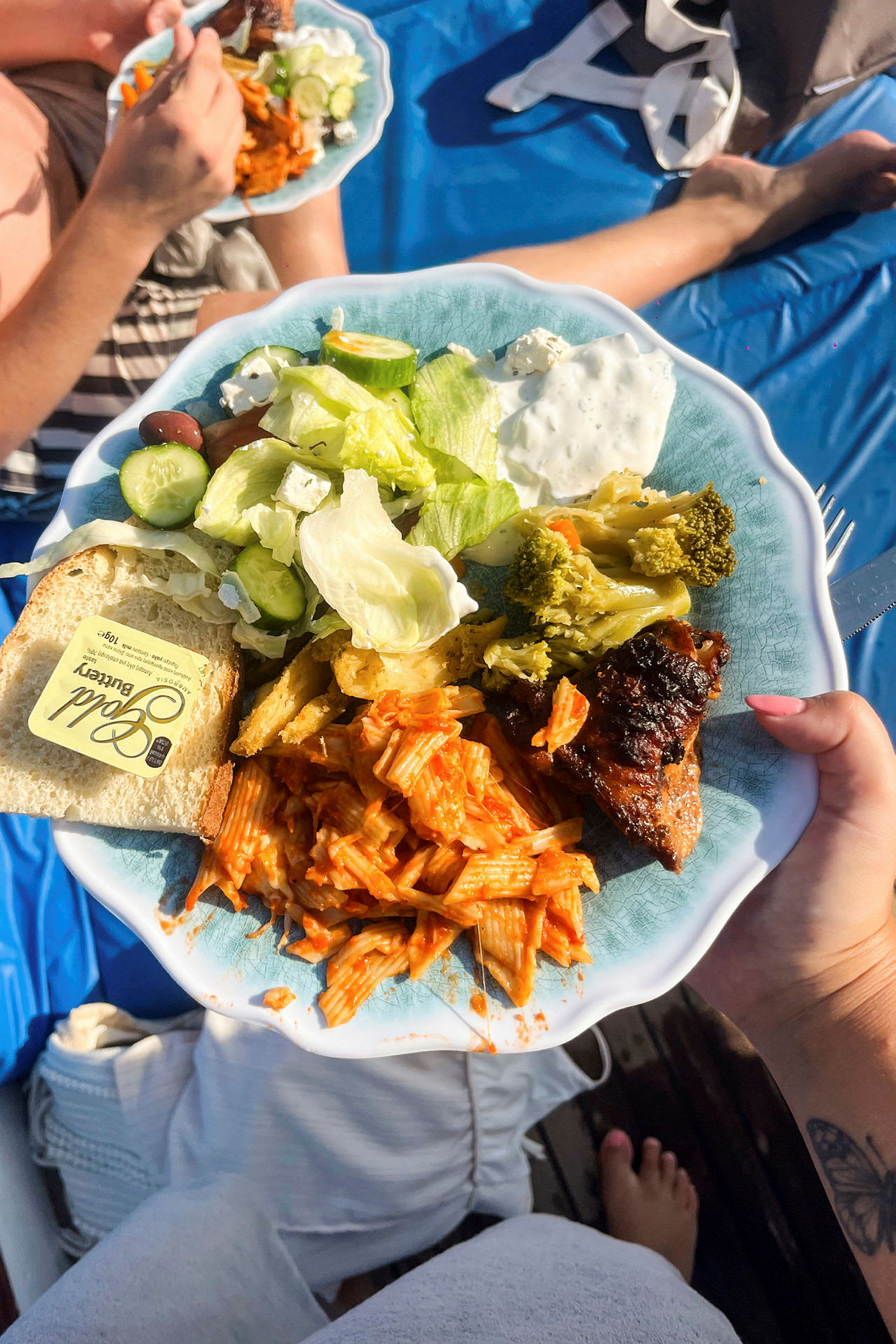 En tallrik med mat innehållande pasta i tomatsås, sallad med grönsaker och fetaost, broccoli, köttbit och en bit bröd med smör. En annan tallrik syns i bakgrunden, samt en person sittande på en blå bordsduk. (AI-genererad text)