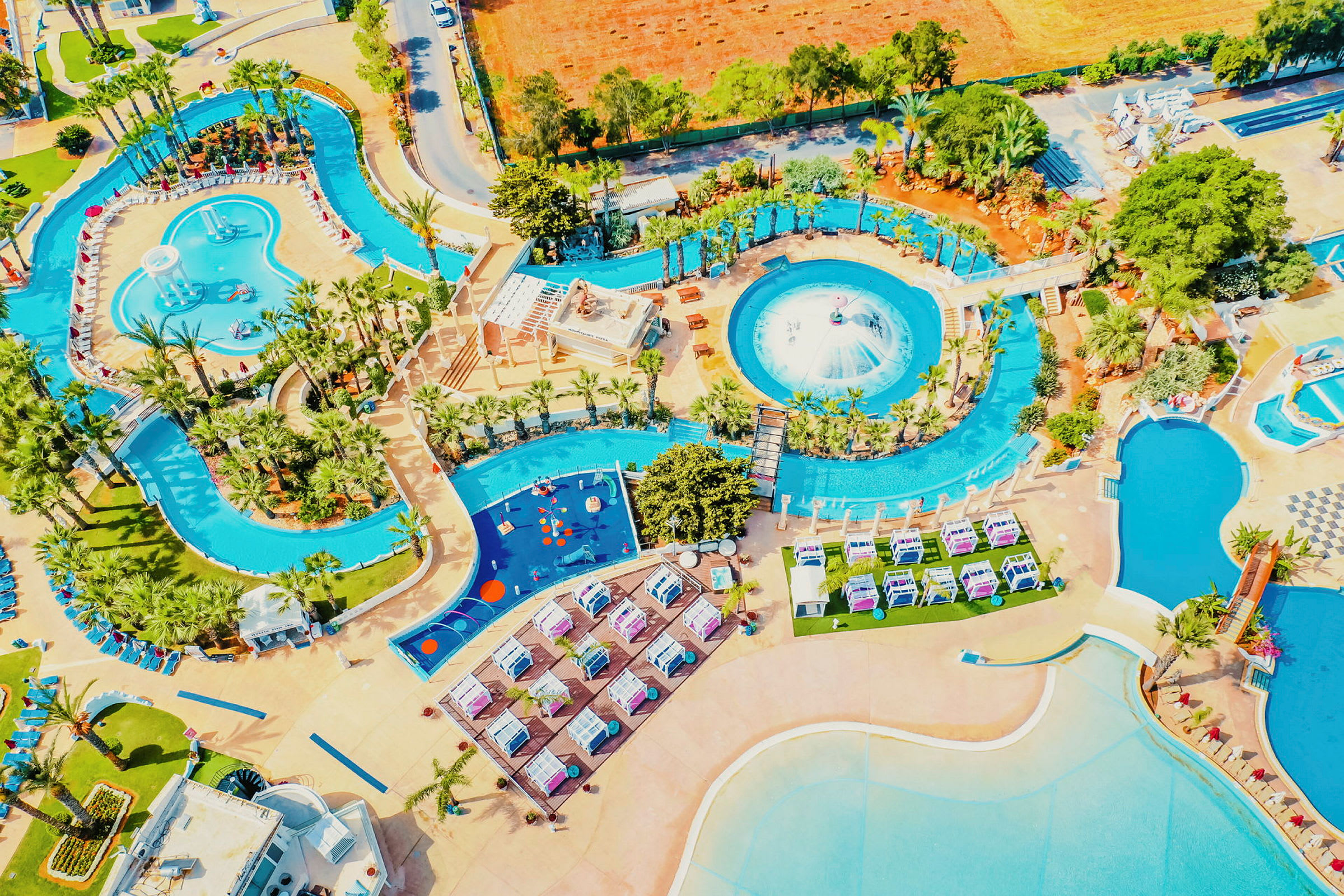 Flygfoto av en färgglad vattenpark med pooler, grönt gräs, solstolar och omgivande grönska. (AI-genererad text)