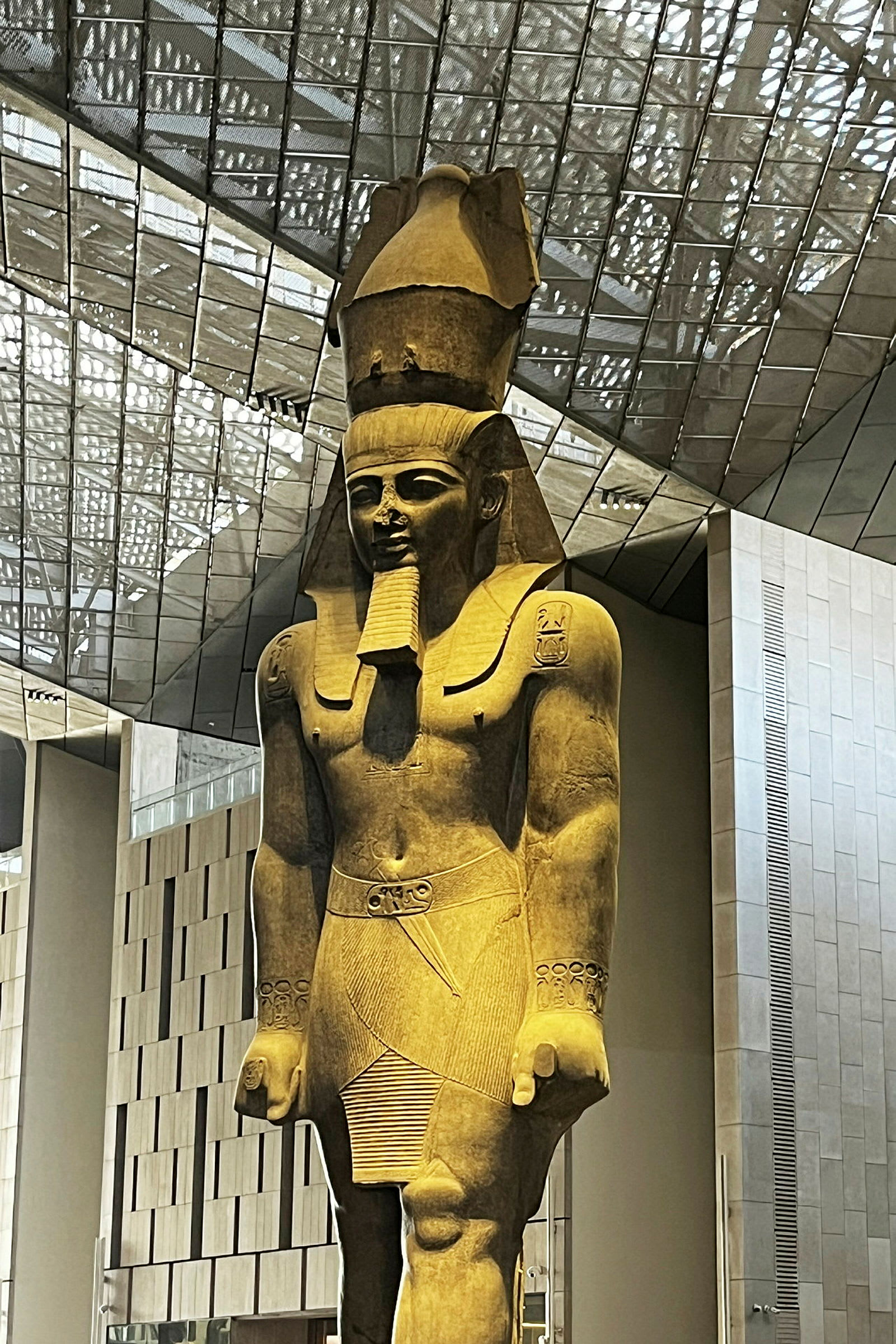 En stor antik staty av en stående mansfigur med detaljerad klädsel och huvudbonad, placerad i en modern byggnad. (AI-genererad text)
