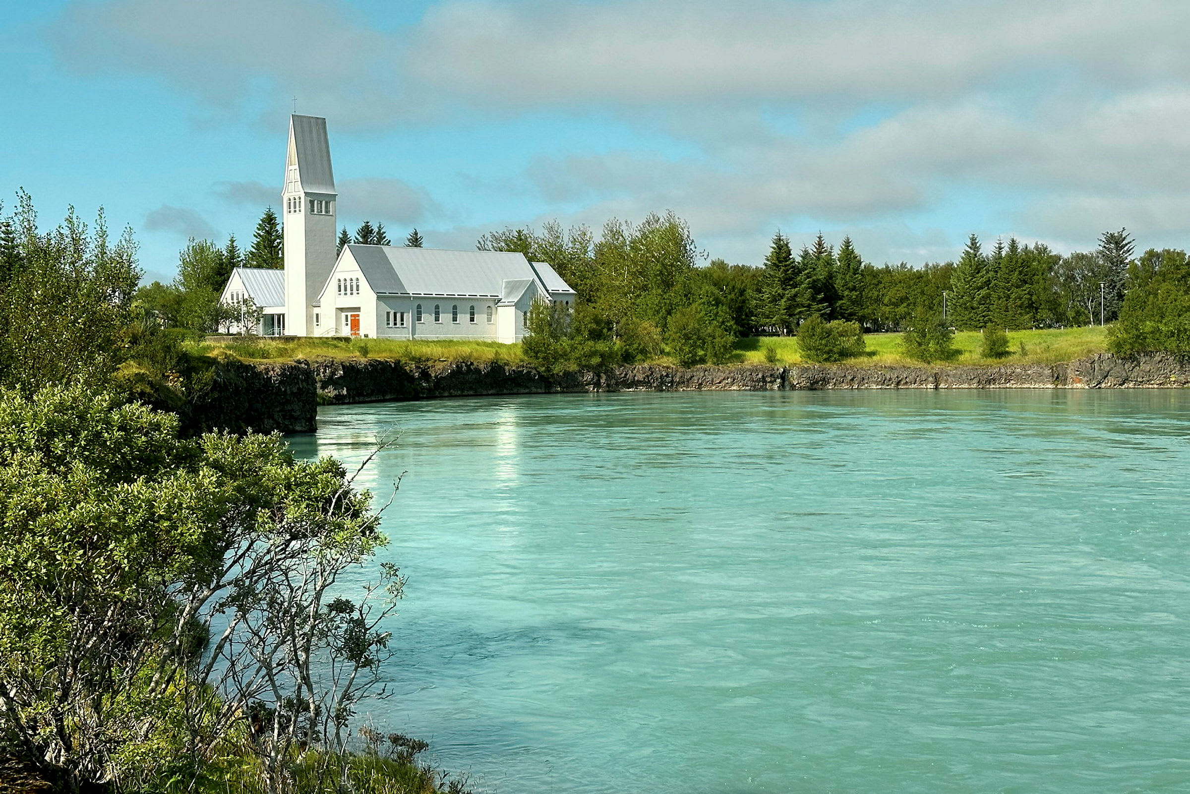 En vit kyrka ligger vid en turkosfärgad flod omgiven av grönska och träd. (AI-genererad text)
