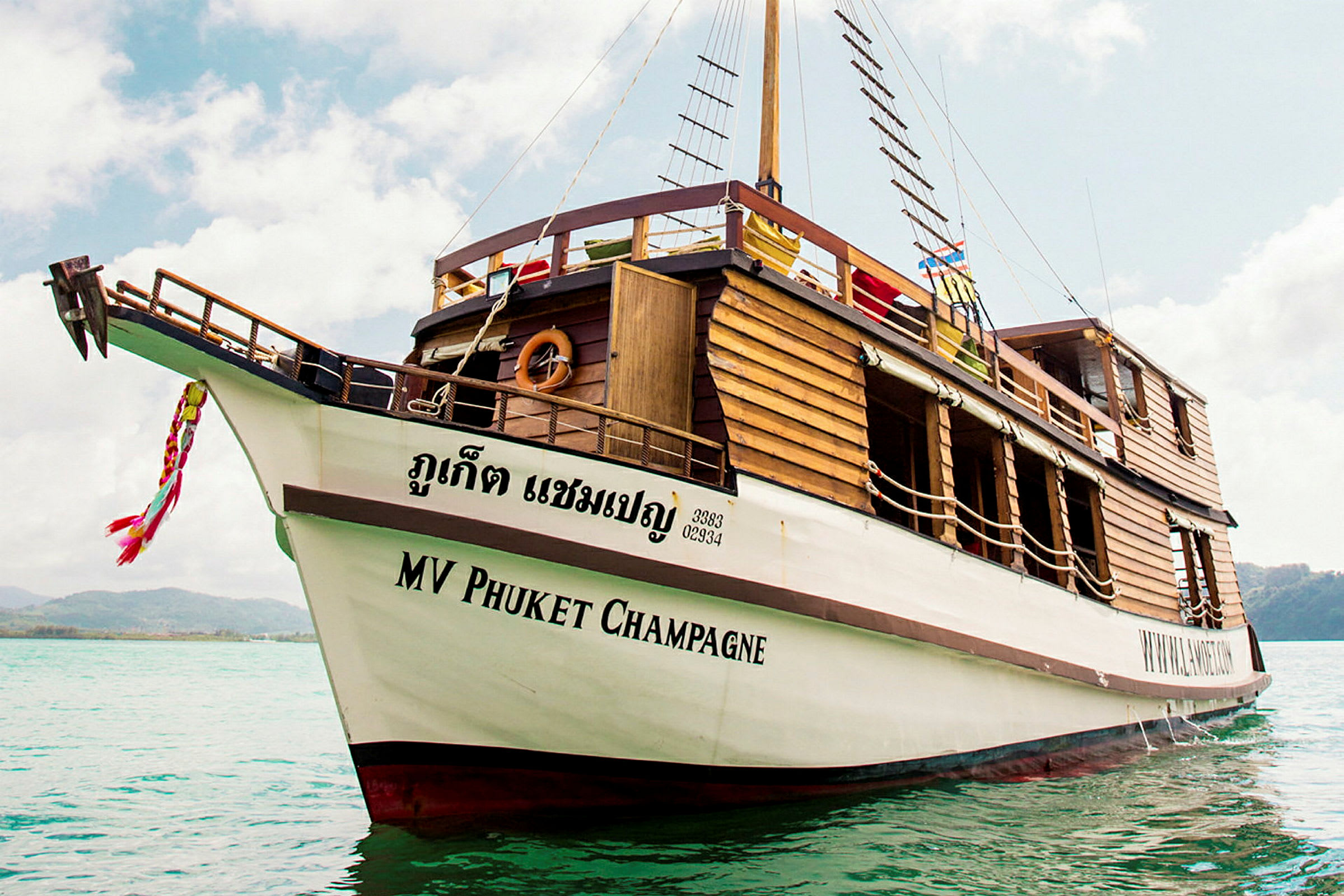 Ett träfartyg med texten 'MV Phuket Champagne' på skrovet ligger förankrat i klart vatten. (AI-genererad text)