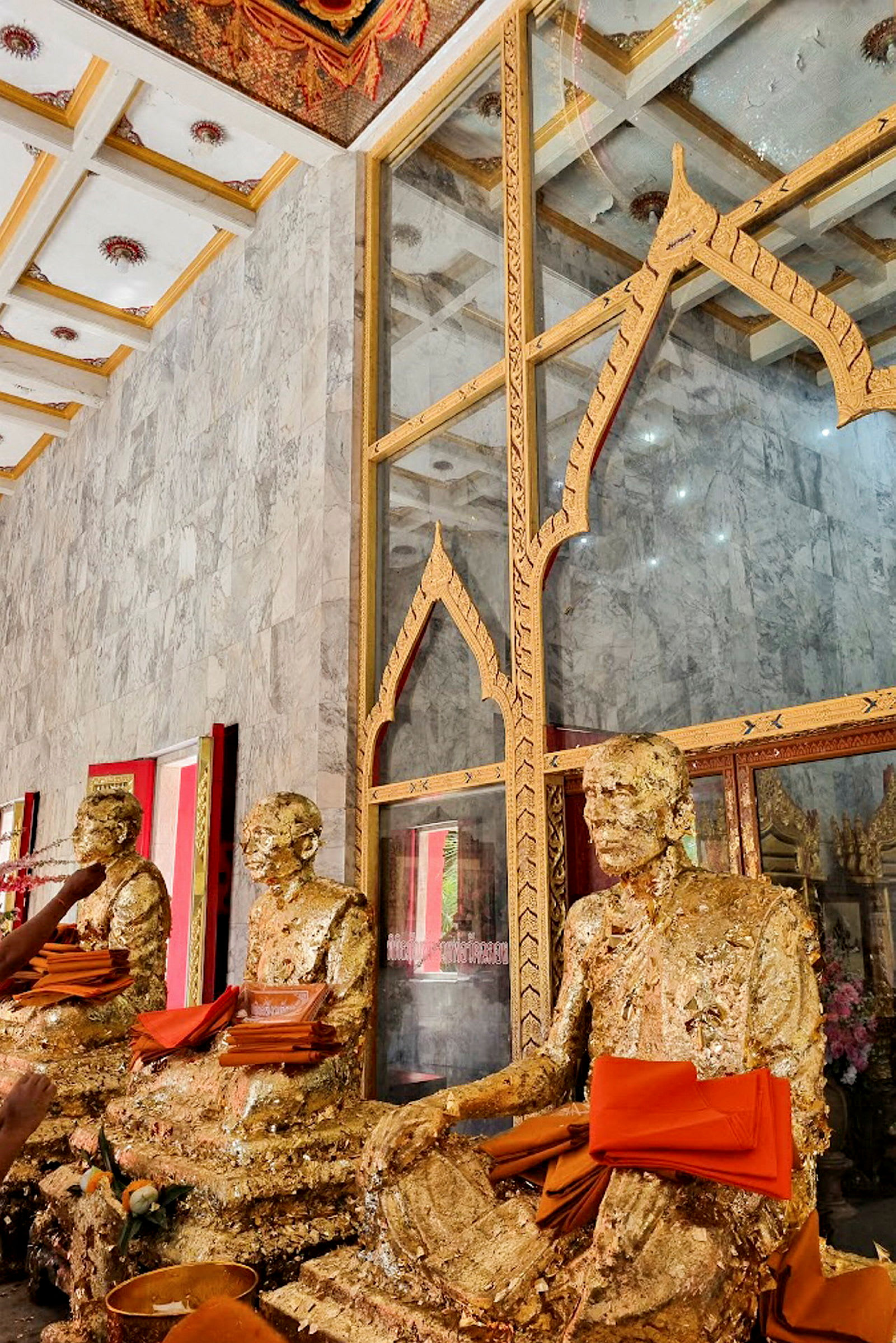 Bild av guldfärgade buddhistiska statyer dekorerade med orange textilier framför en glasvägg med utsmyckningar. (AI-genererad text)