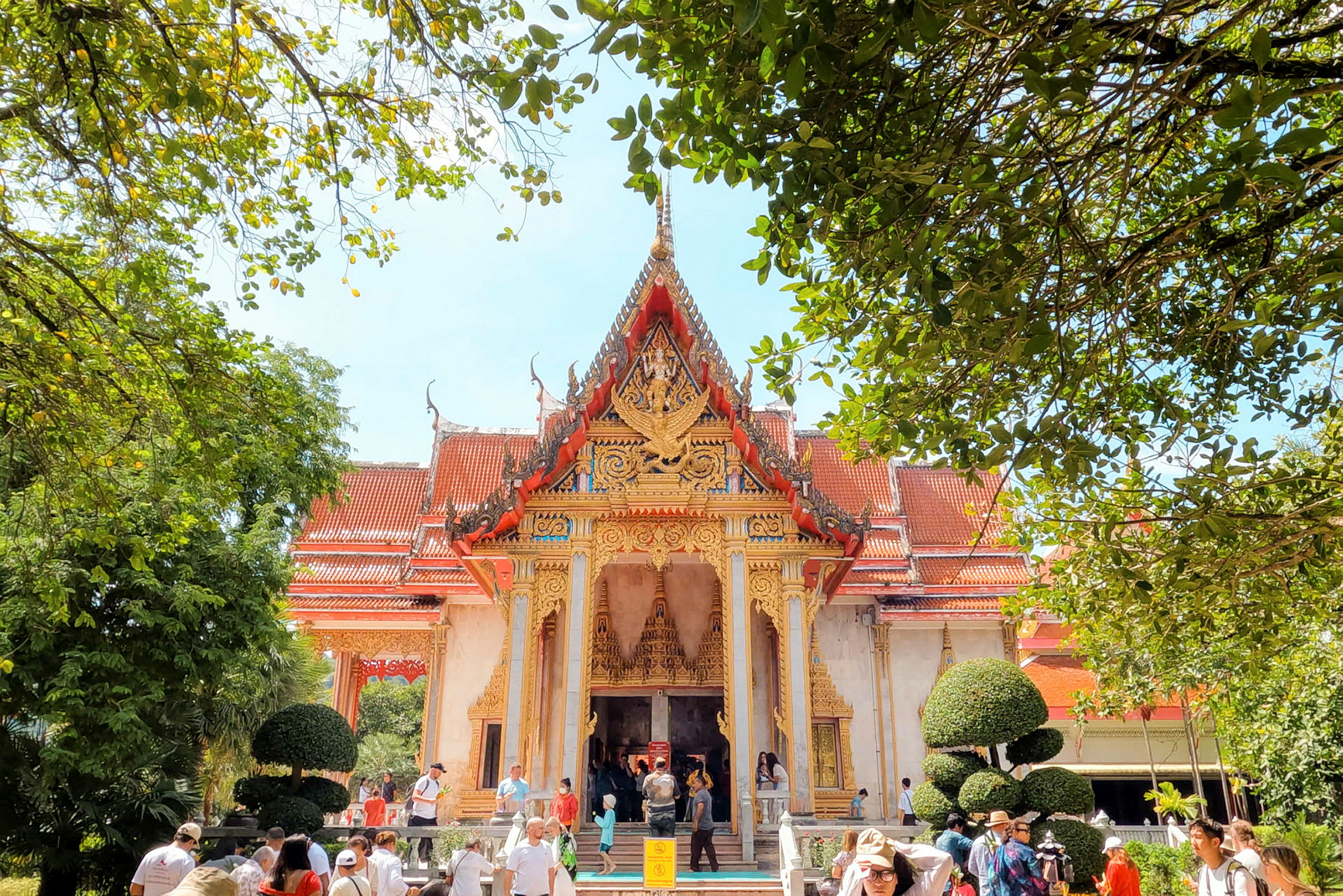 Bild på ett detaljrikt thailändskt tempel omgivet av frodig vegetation, med besökare framför byggnaden. (AI-genererad text)