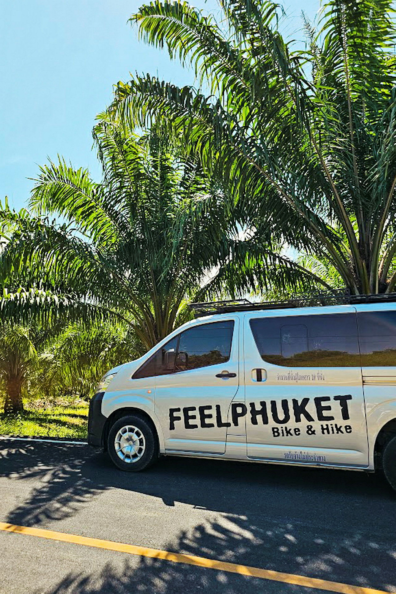 En minibuss med texten 'Feel Phuket Bike & Hike' parkerad vid sidan av vägen nära palmer. (AI-genererad text)