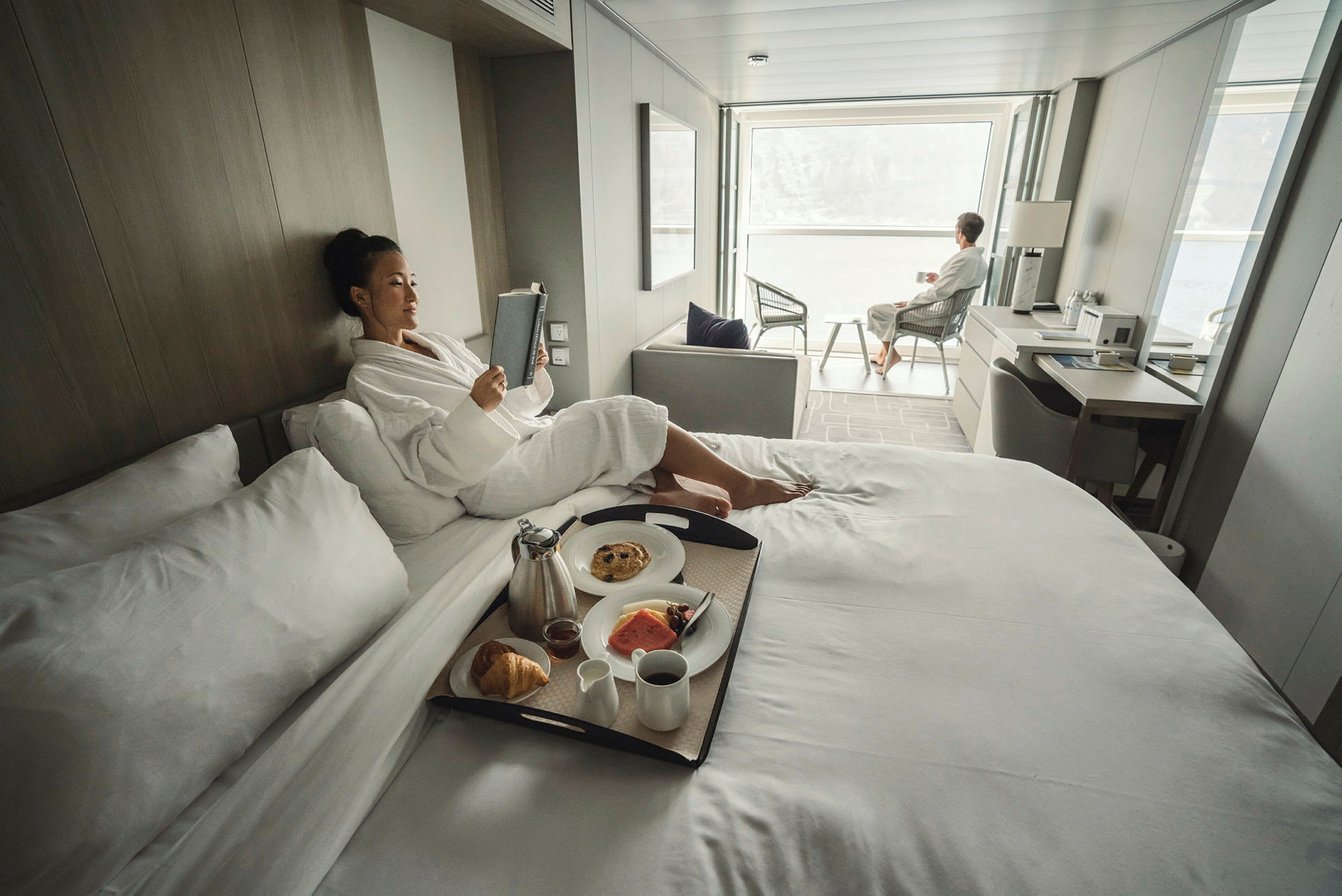 Personer kopplar av i ett ljust och modernt hotellrum, med frukost serverad på sängen och sittplats vid en stor fönstervy. (AI-genererad text)