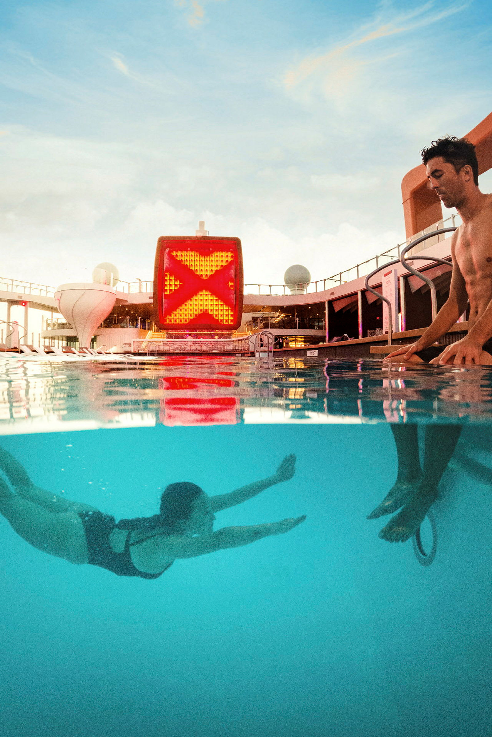 En person simmar i en pool under vatten medan en annan står vid poolkanten. Bakgrunden visar ett fartygs öppna däck och himlen. (AI-genererad text)