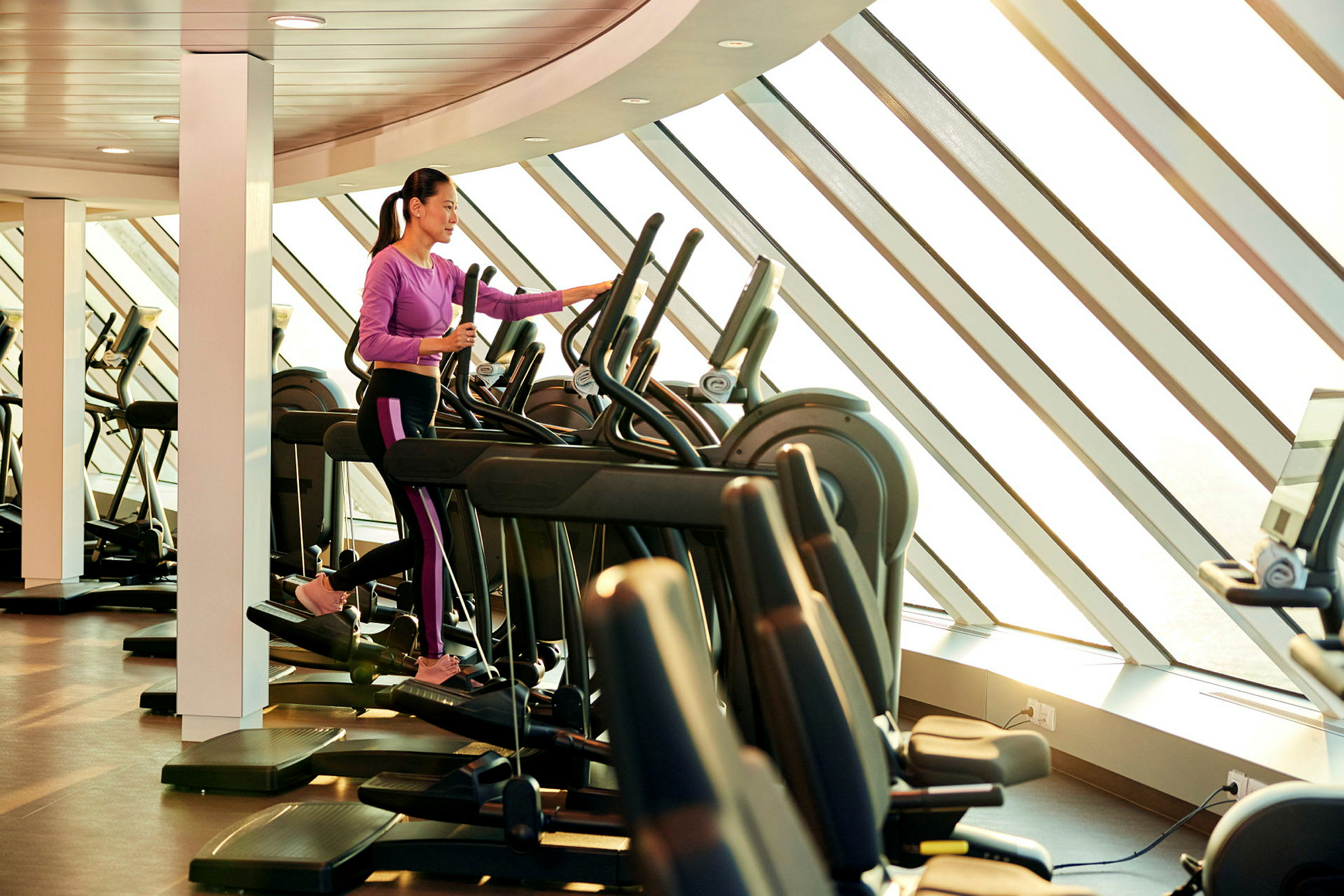 Person tränar på en elliptisk träningsmaskin i ett gym med stora fönster. (AI-genererad text)