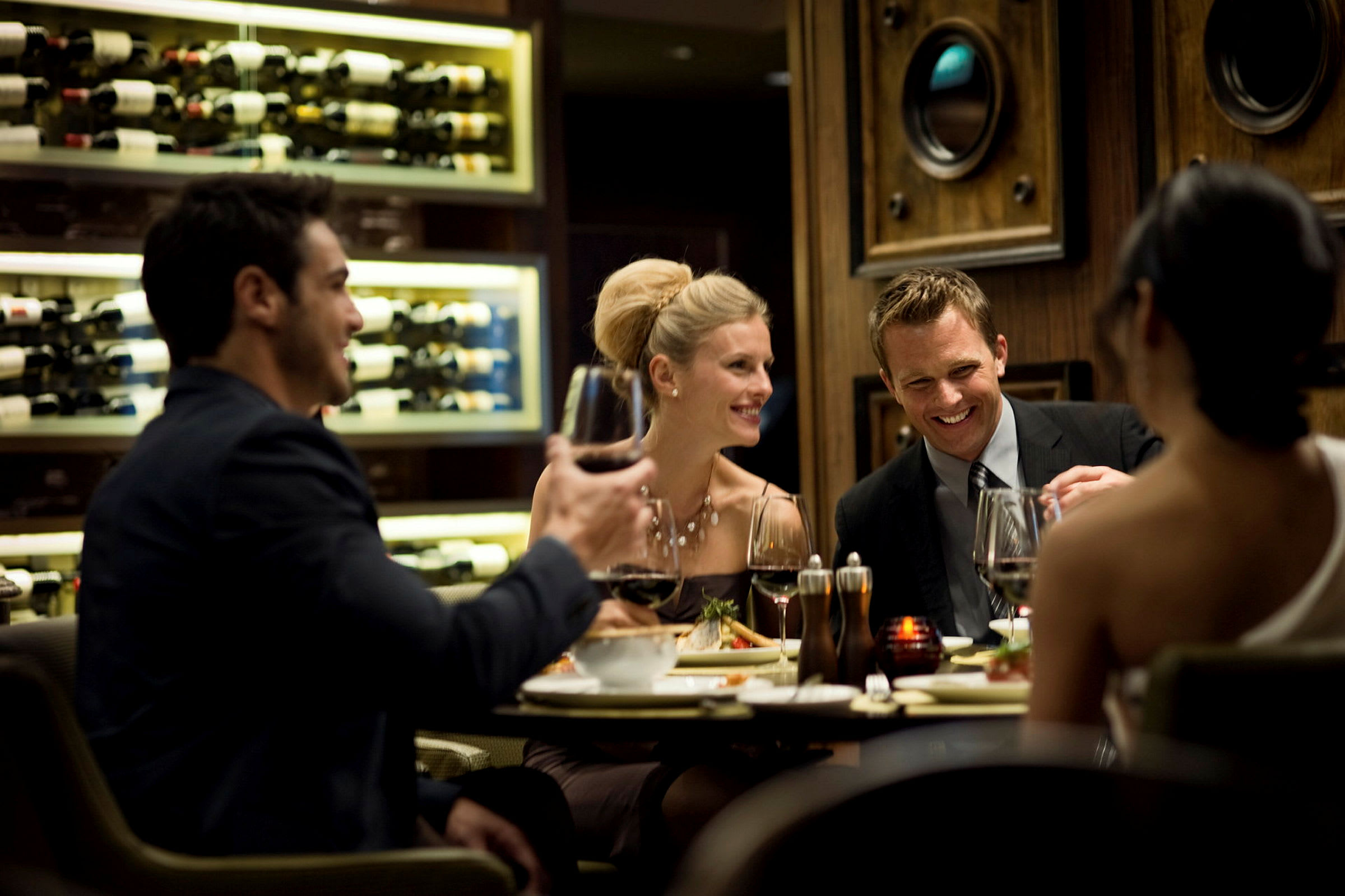 En grupp människor njuter av mat och vin vid ett bord i en elegant restaurang med en vägg fylld av vintillgångar i bakgrunden. (AI-genererad text)