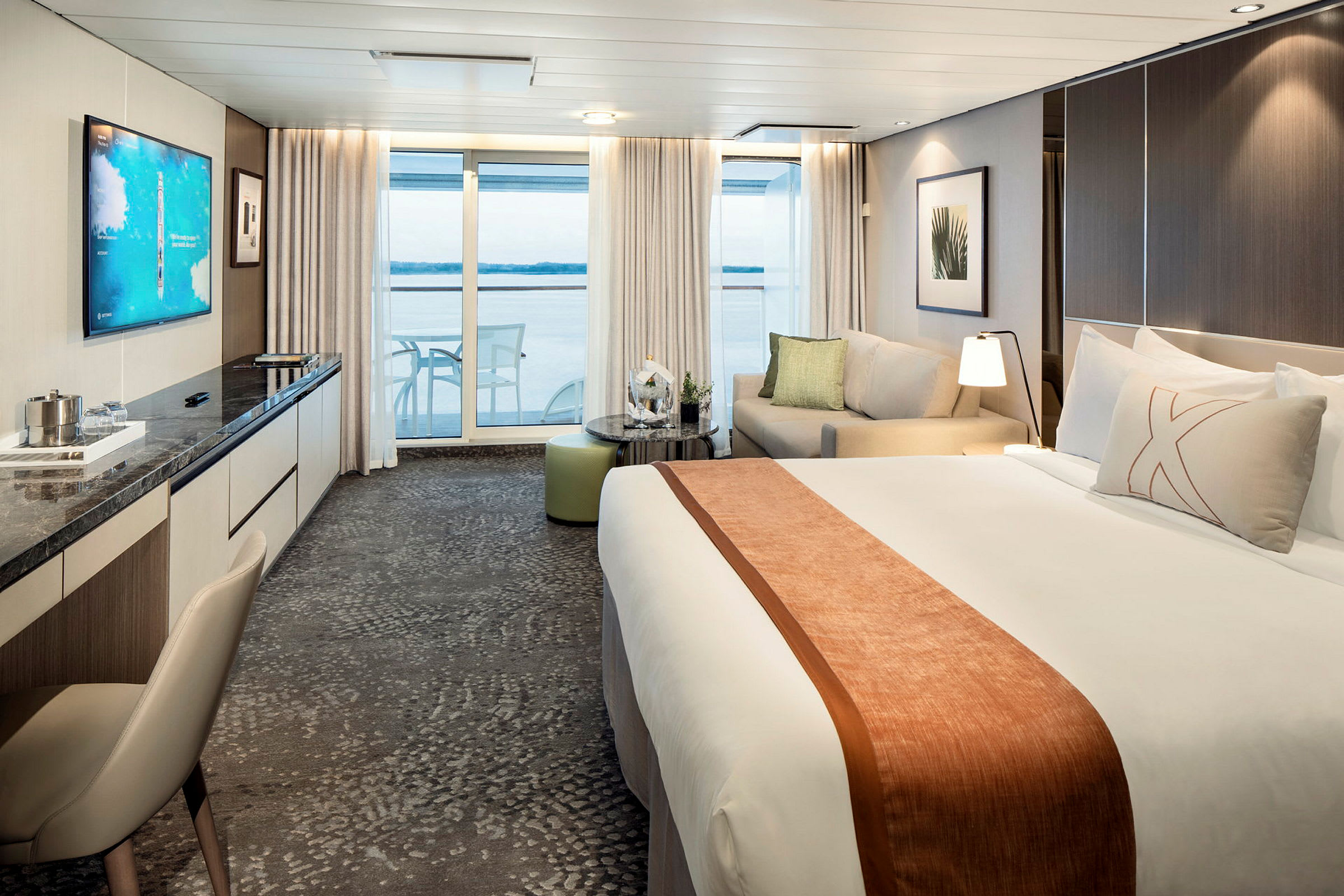 Ett modernt hotellrum med en stor säng, soffa och ett bord, samt utsikt genom glasdörrar mot en balkong och havet. (AI-genererad text)