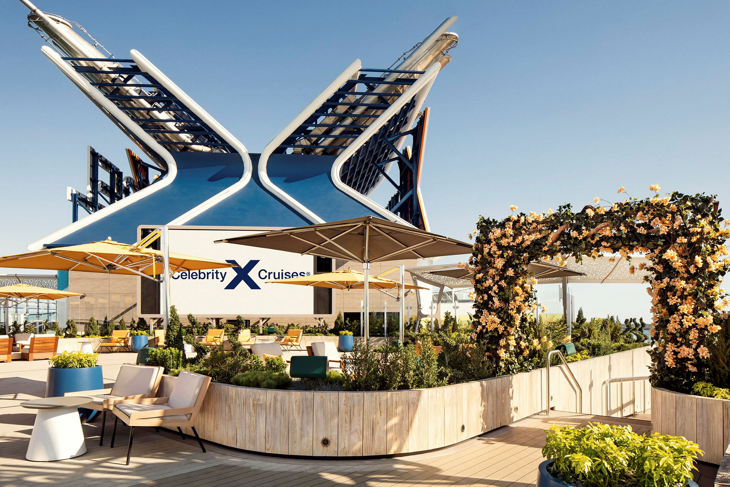 Ett fartygsdäck med sittgrupper, gula parasoller och välskött grönska, med logotypen 'Celebrity Cruises' synlig. (AI-genererad text)