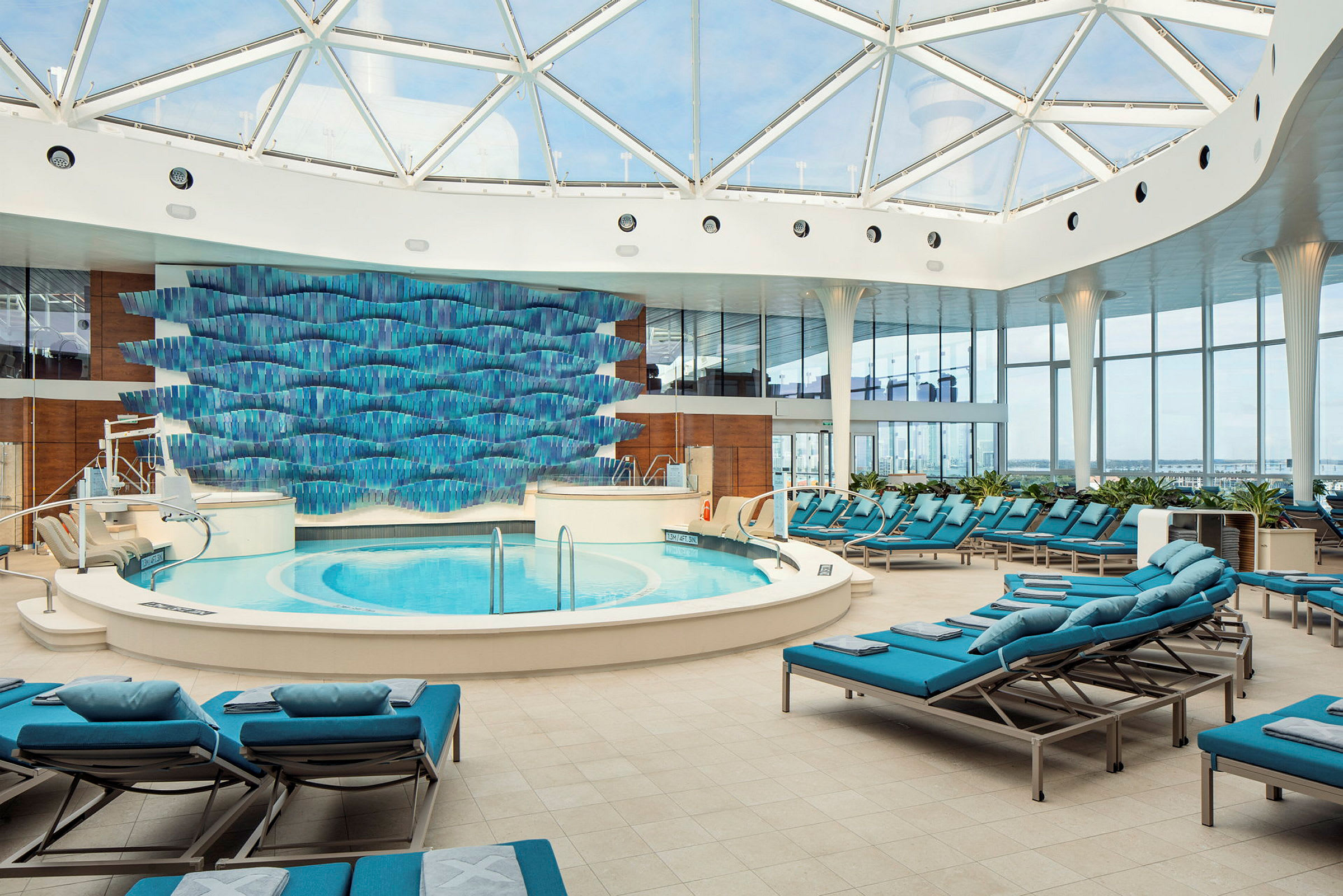 Ett ljust inomhuspoolområde med en rund pool, solstolar och en dekorerad vägg med blå konstverk. (AI-genererad text)