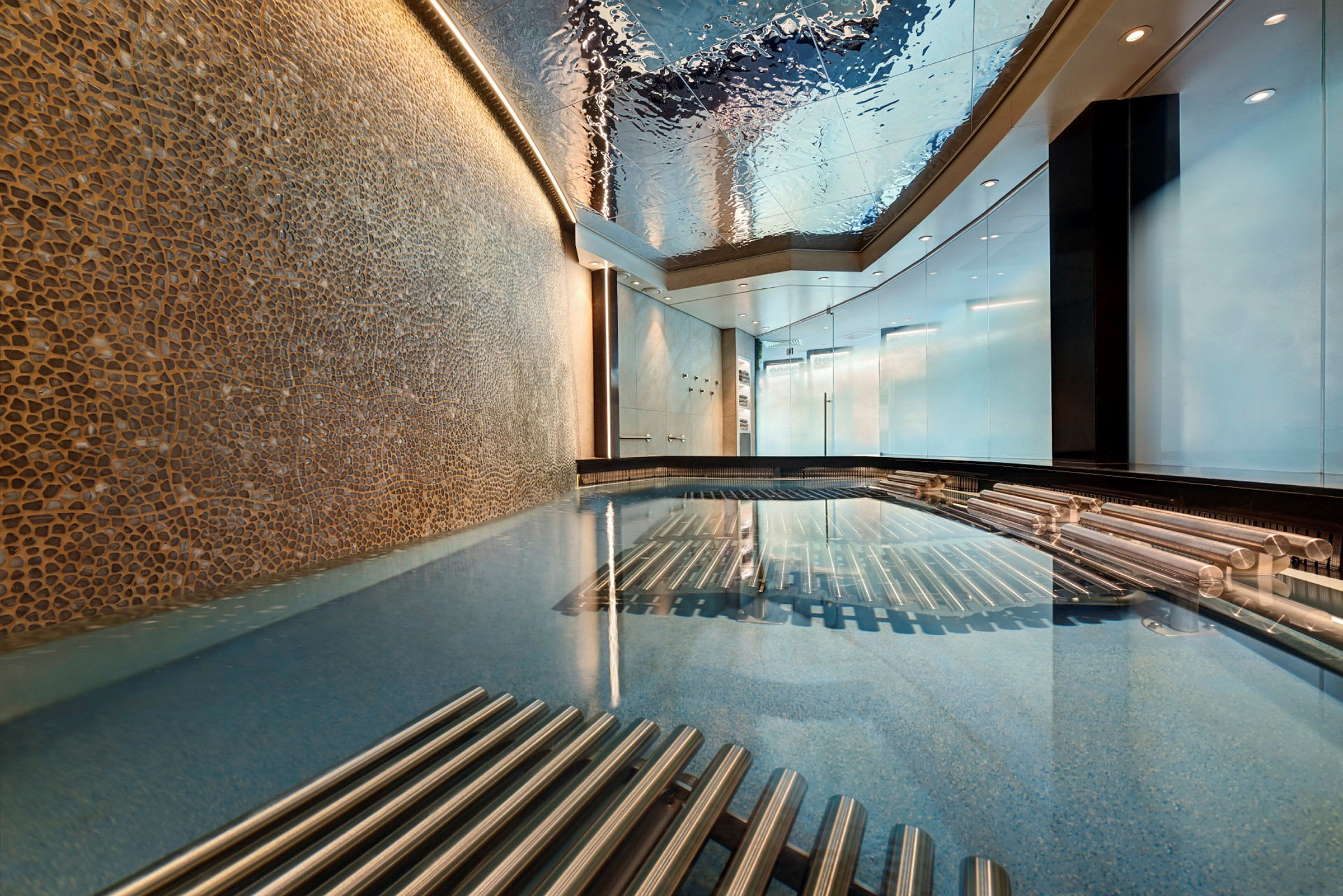 En inomhuspool med ett tak som reflekterar ljus och omgivning, flankerad av metalliska detaljer och väggar med dekorativa mosaiker. (AI-genererad text)