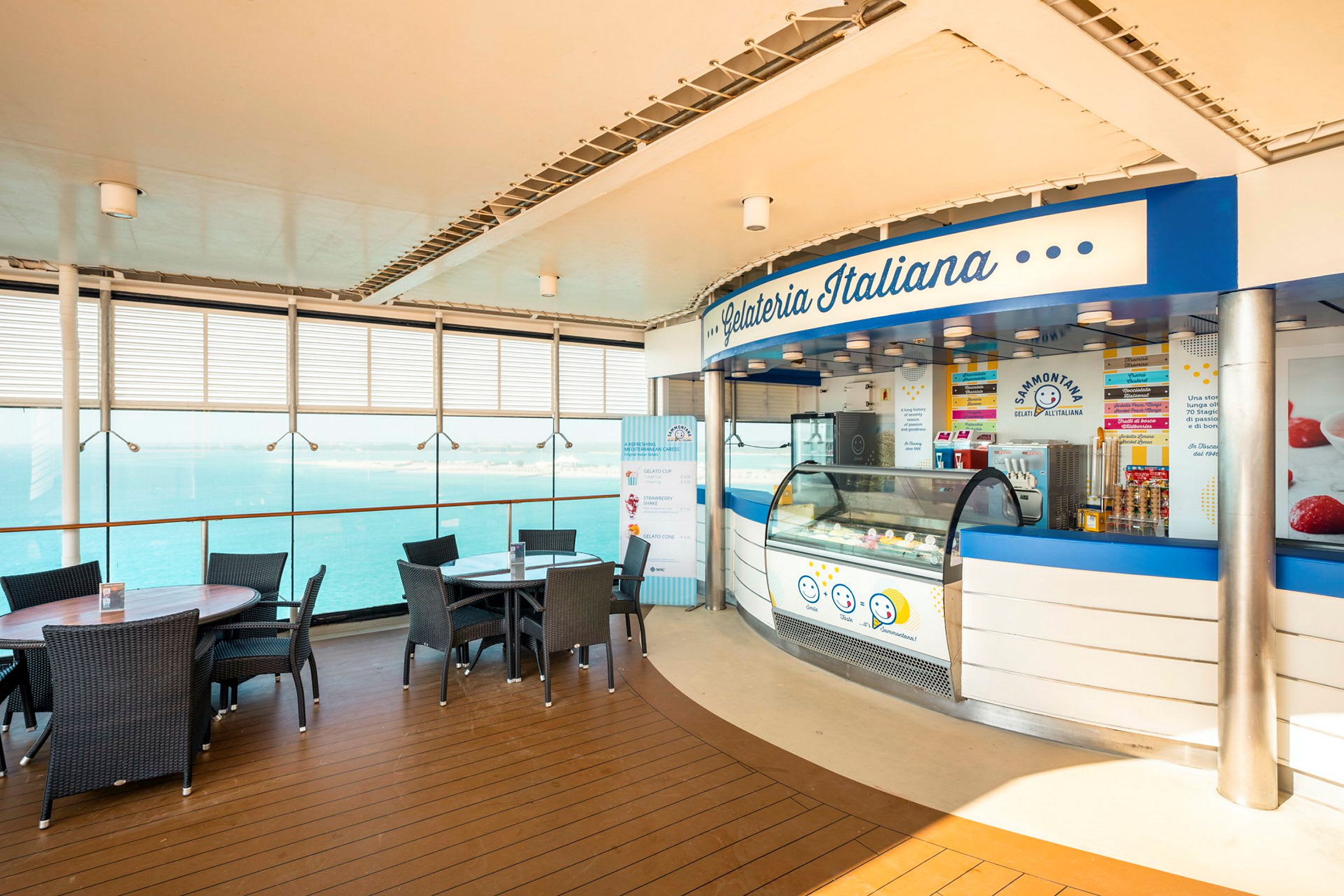En glasskiosk med namnet Gelateria Italiana belägen vid ett område med bord och stolar nära glasfönster med utsikt över havet. (AI-genererad text)