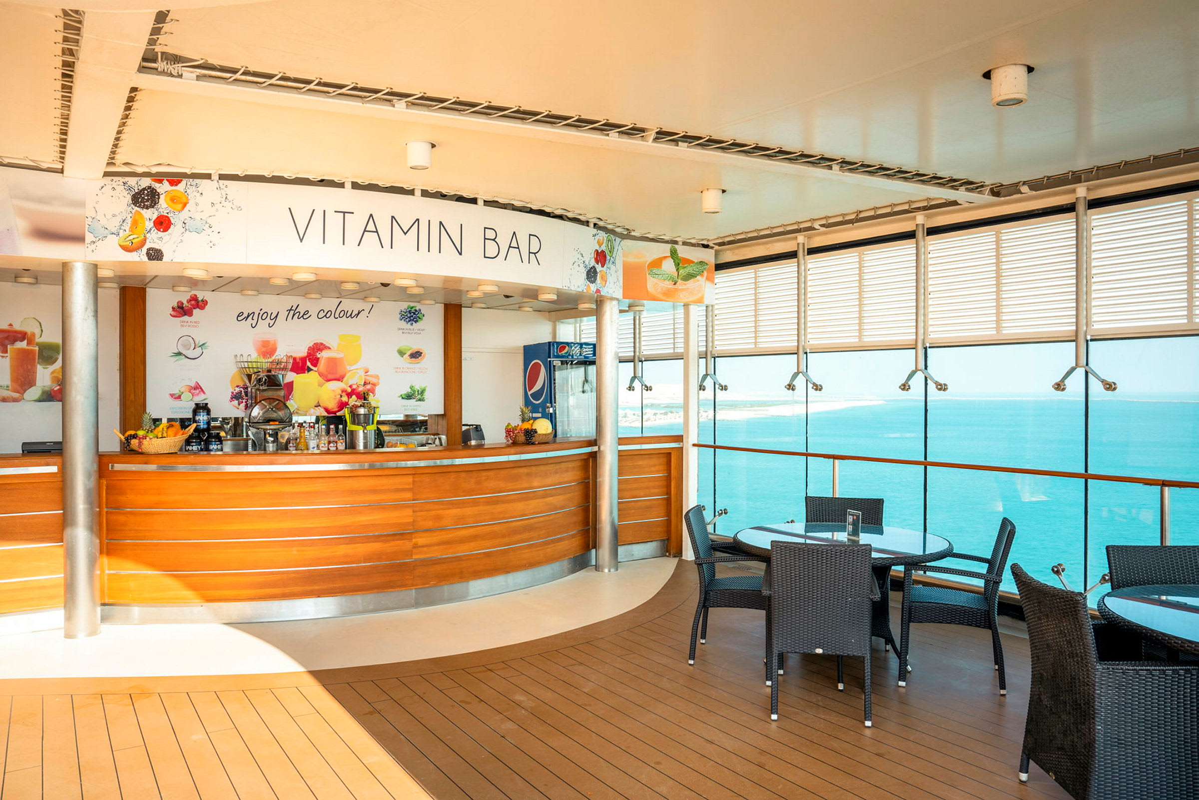Bild av en vitaminbar med träpaneler, färgglada frukter som dekoration och en glasvägg som vetter mot vattnet. Några bord med glasskivor och mörka rottingstolar finns i förgrunden. (AI-genererad text)