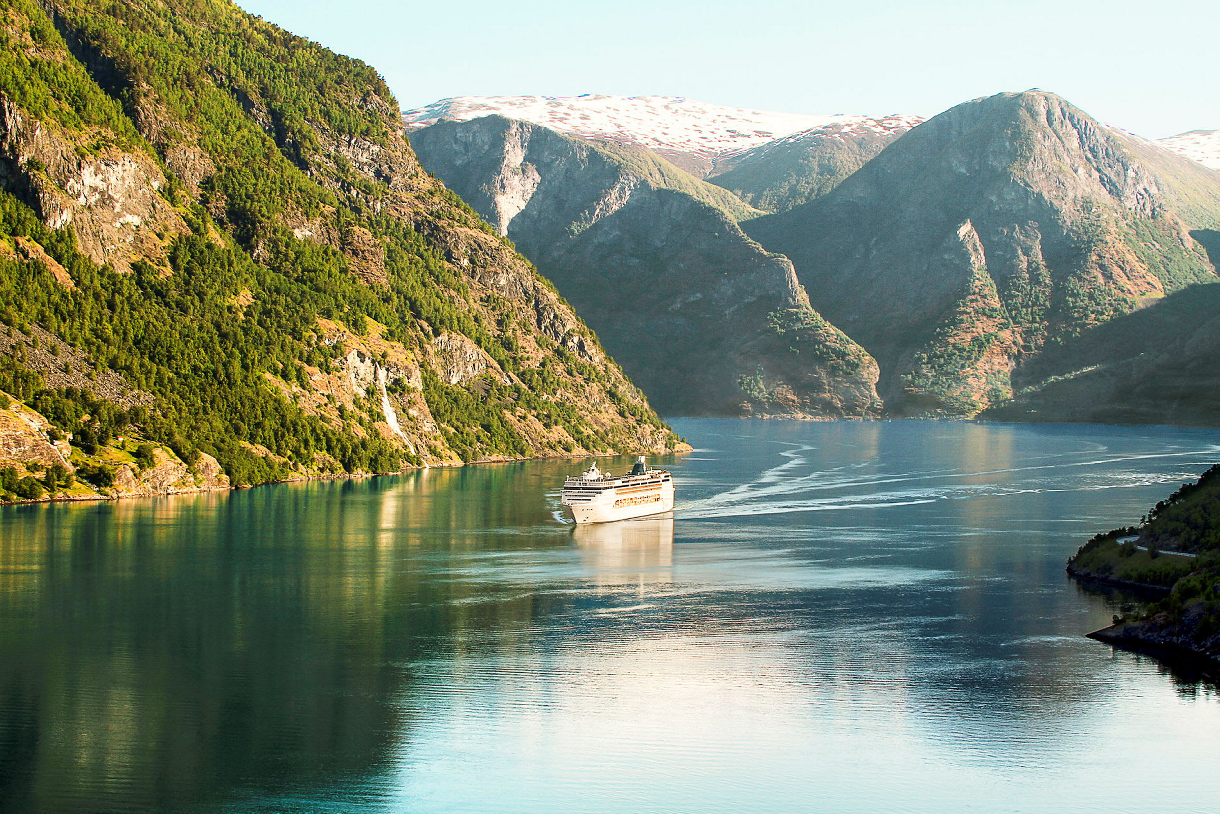 Bild på en kryssningsfartyg som seglar genom en fjord med höga bergsklädda sluttningar och klart vatten. (AI-genererad text)