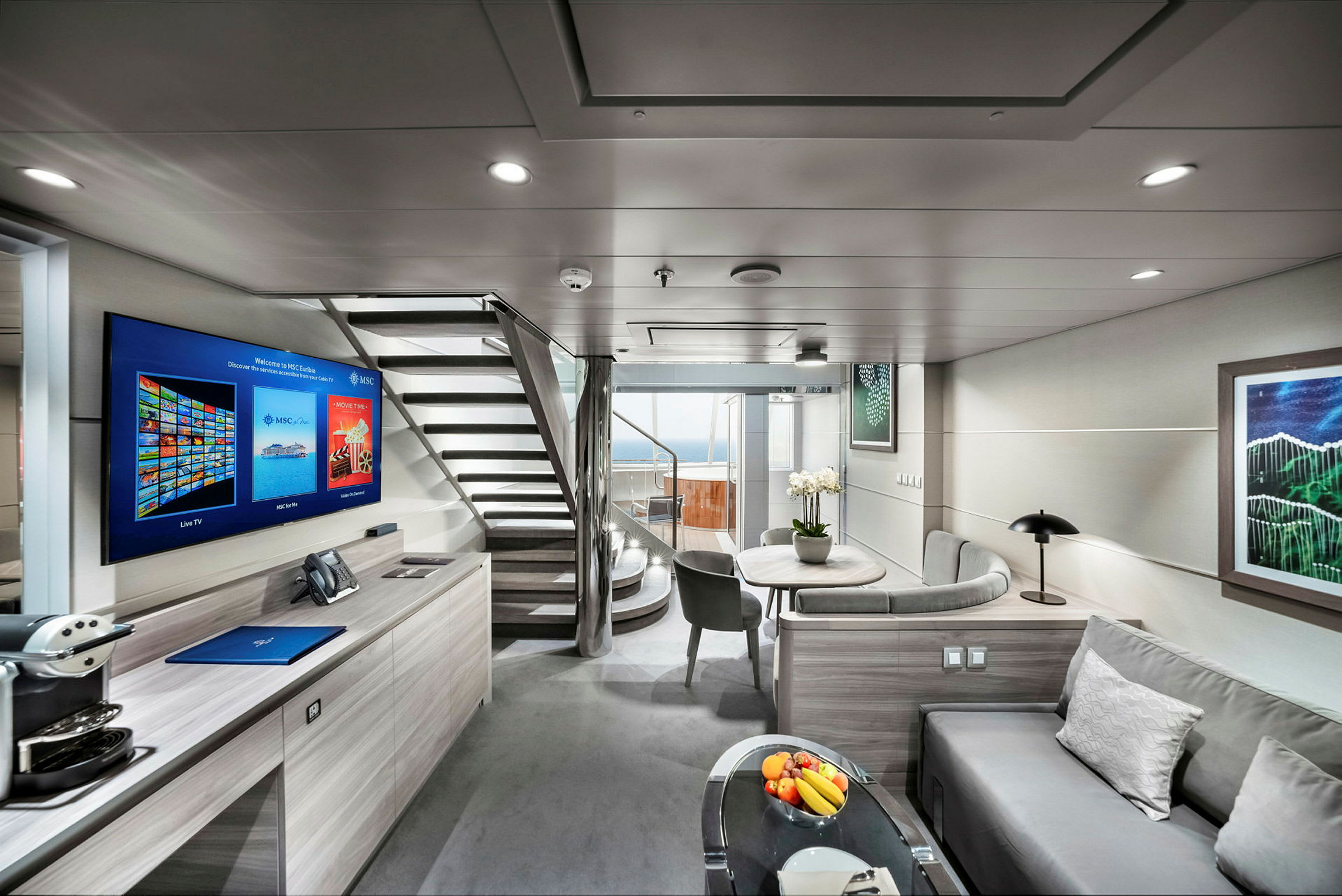 En modern och ljus interiör av en hotellsvit med soffa, bord, platt-TV och trappa som leder till en övre våning samt en balkong med utsikt. (AI-genererad text)