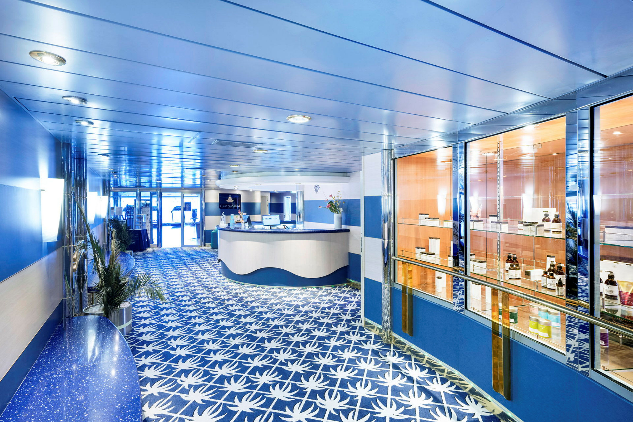 Hotellanläggning med reception och en glasdisplay från spaområdet. Inredningen har ett blått tema med palmbladsmönster på golvet. (AI-genererad text)