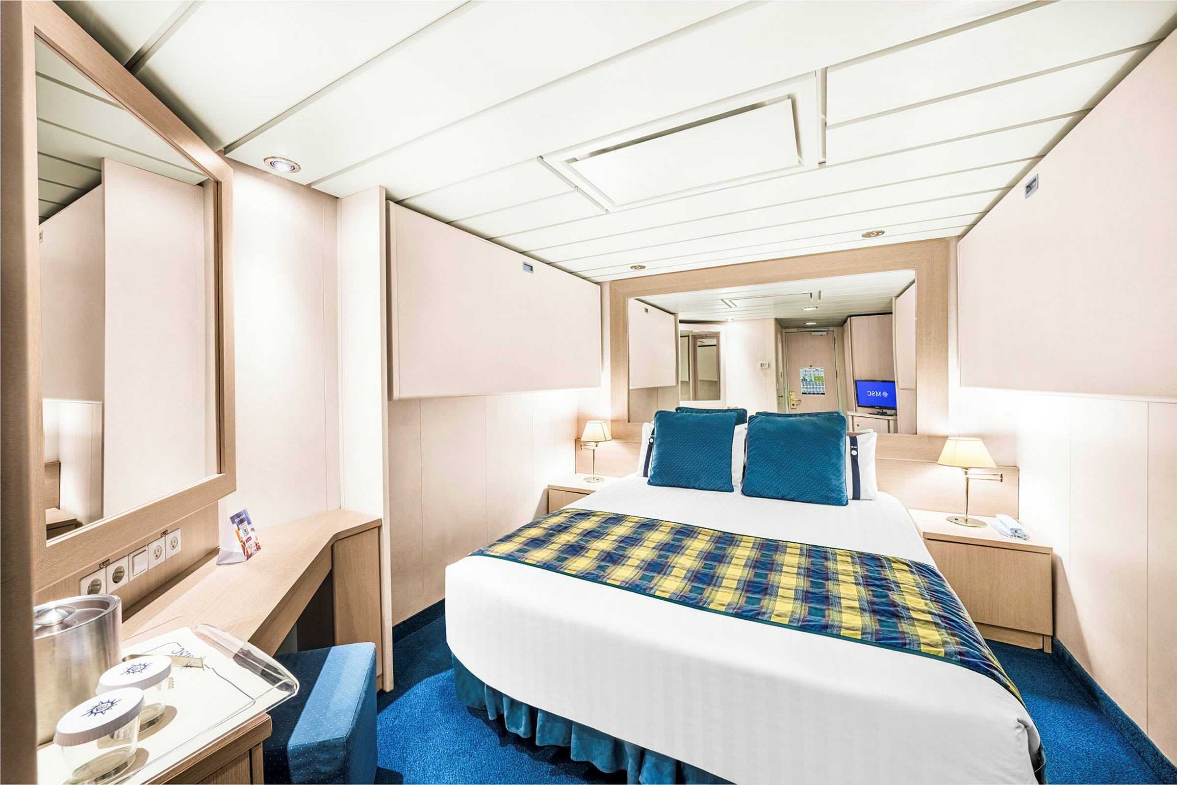 En bild av ett hotellrum med en modern inredning, en säng med blå kuddar och guldbetonat täcke, två nattduksbord och speglar. (AI-genererad text)