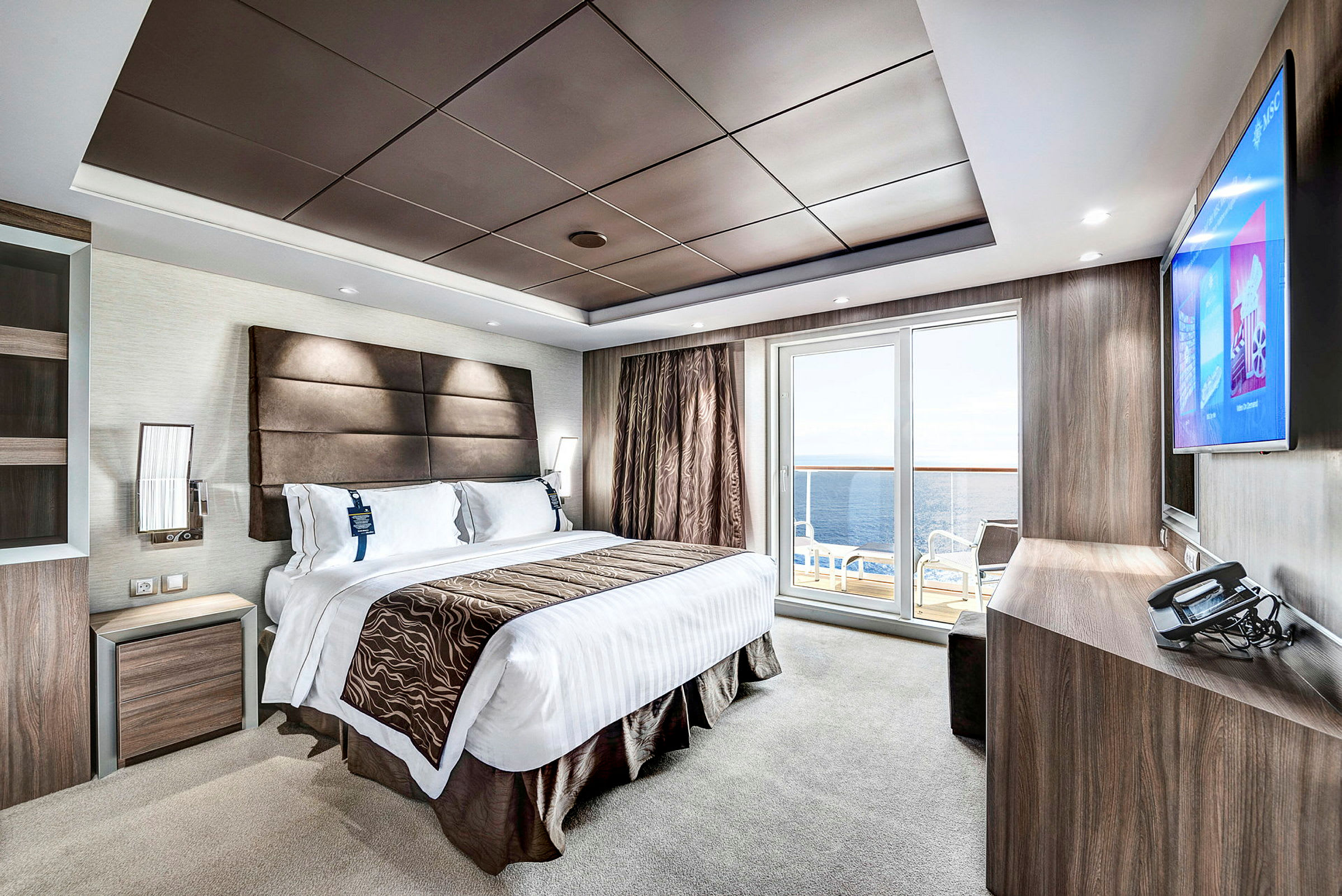 Ett modernt hotellrum med en stor säng, en platt-TV, ett skrivbord och en balkong med utsikt mot havet. (AI-genererad text)