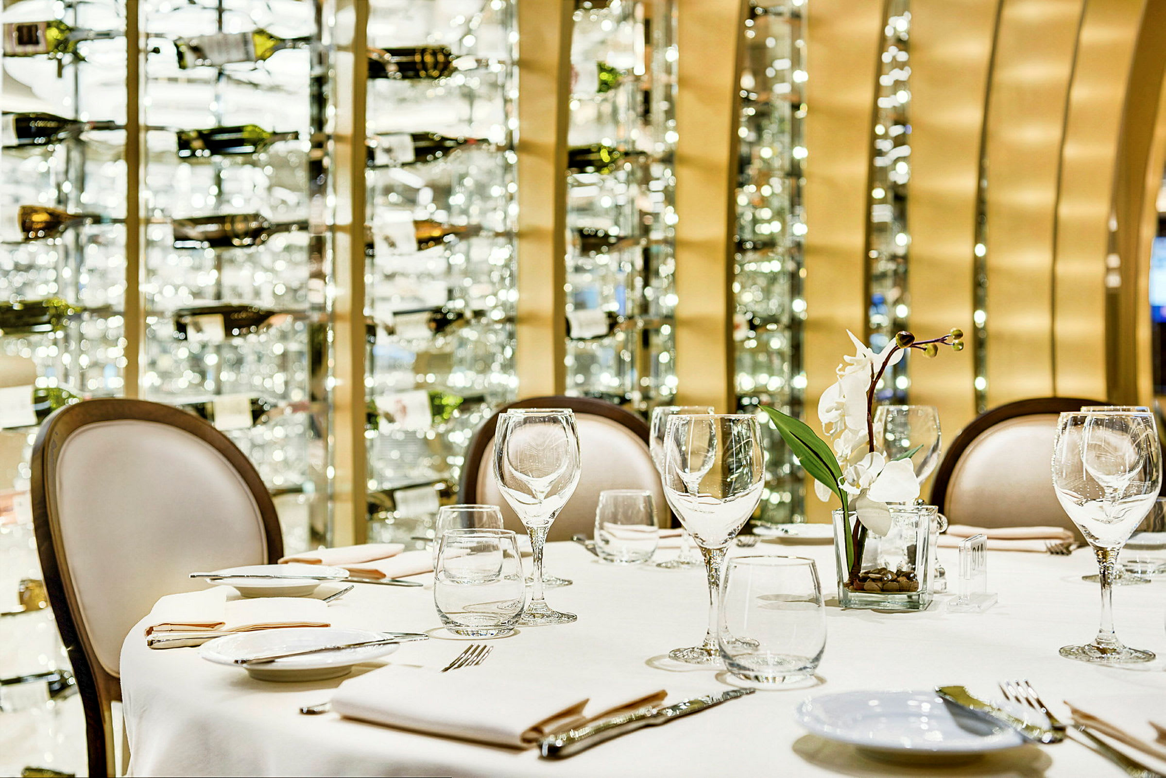 Dukning med glas, tallrikar och en vit blomma på en restaurang med vinflaskor i bakgrunden. (AI-genererad text)